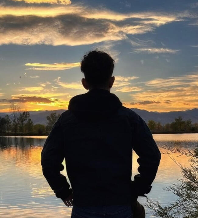Poet Andrew Gibson, source: https://www.instagram.com/p/DJXFHUvutHg/?img_index=17 Andrew Gibson stands silhouetted, facing away from us, in front of a sunset view over a body of water with trees and mountains in the background.